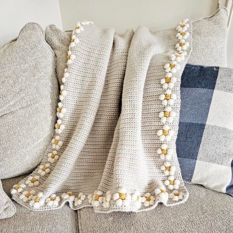 May include: A crocheted throw blanket in a light beige color with a white and yellow daisy border.