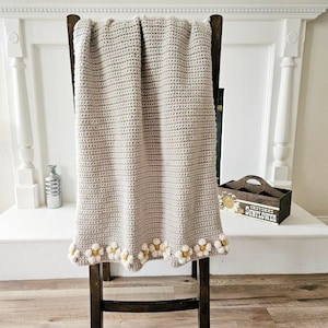 May include: A crocheted throw blanket in a light beige color with a white and yellow daisy trim. The blanket is draped over a wooden chair.