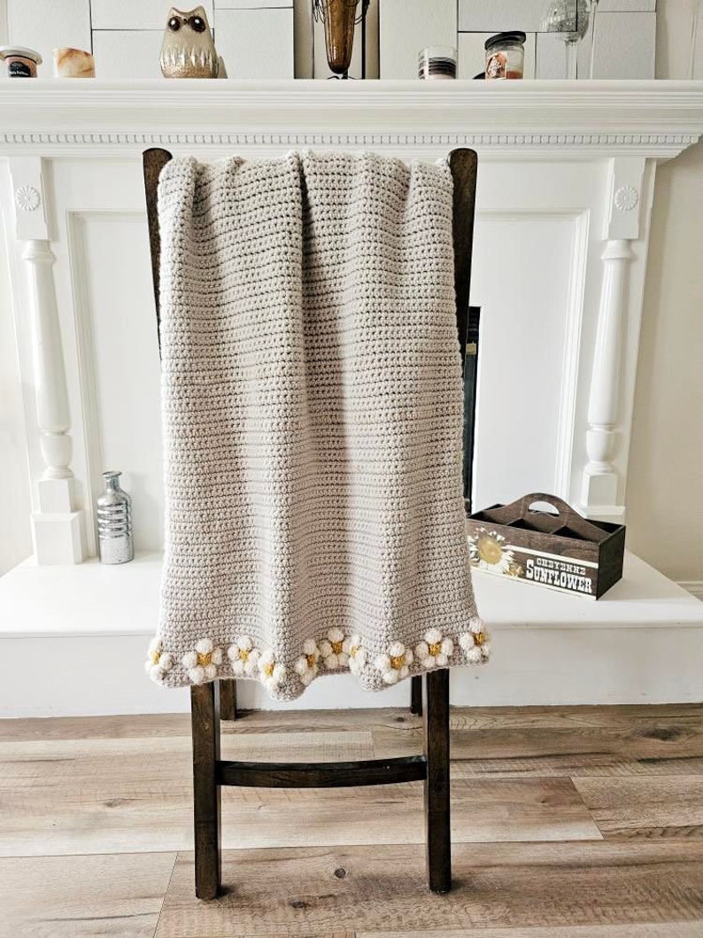 May include: A crocheted throw blanket in a light beige color with a white and yellow daisy trim. The blanket is draped over a wooden chair.