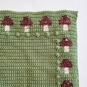 May include: A close-up of a crocheted blanket with a green background and a pattern of burgundy and white mushrooms. The mushrooms are evenly spaced across the top and along the right side of the blanket. The blanket has a textured appearance.
