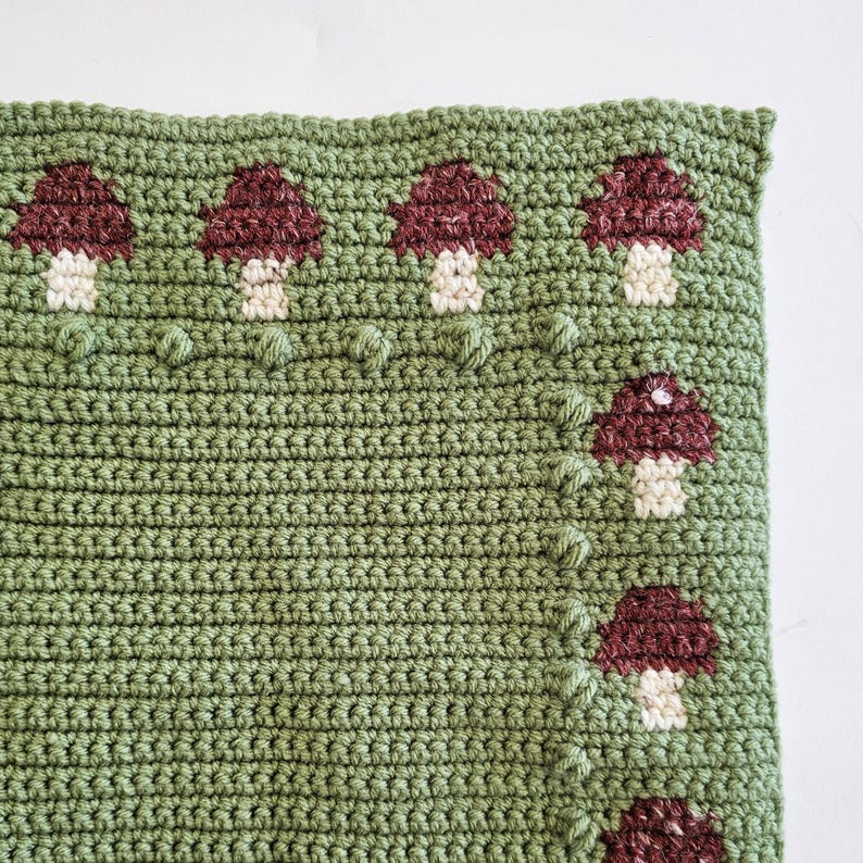 May include: A close-up of a crocheted blanket with a green background and a pattern of burgundy and white mushrooms. The mushrooms are evenly spaced across the top and along the right side of the blanket. The blanket has a textured appearance.