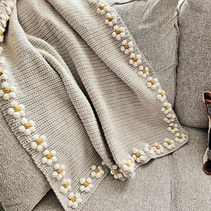 May include: A gray crocheted throw blanket with a white daisy border. The blanket is folded and draped over a gray couch.