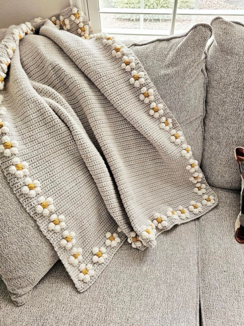 May include: A gray crocheted throw blanket with a white daisy border. The blanket is folded and draped over a gray couch.