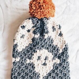 May include: A gray and white crocheted beanie with a large orange pom pom on top.