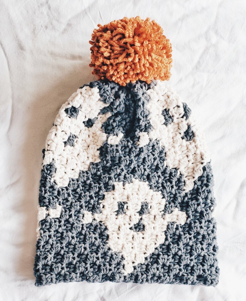 May include: A gray and white crocheted beanie with a large orange pom pom on top.