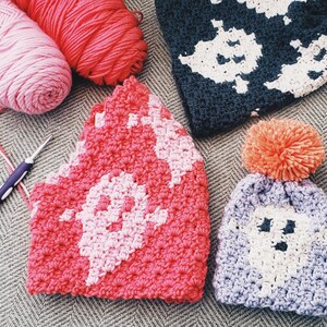May include: Three crocheted hats with ghost designs. One hat is pink and white, one is purple and white, and one is dark blue and white. The hats are on a gray and white patterned surface.
