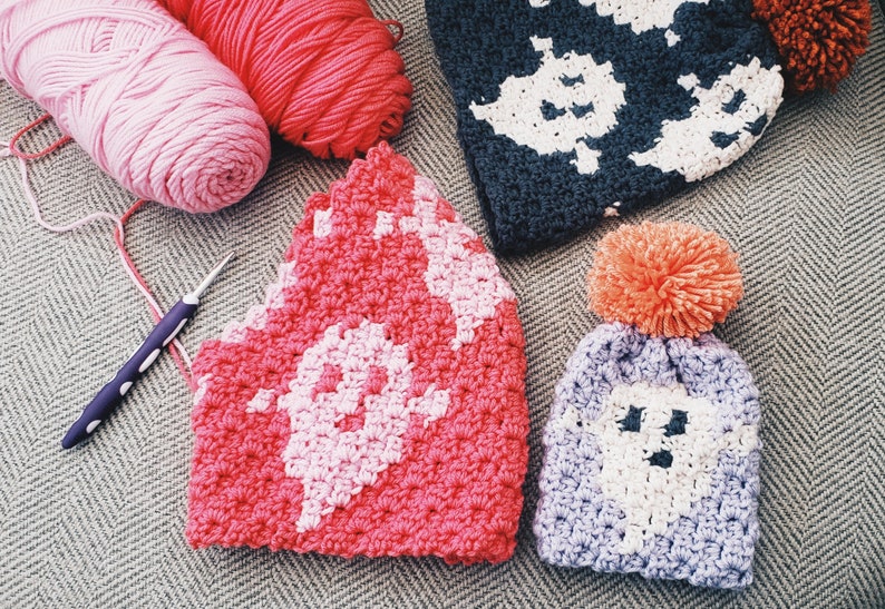 May include: Three crocheted hats with ghost designs. One hat is pink and white, one is purple and white, and one is dark blue and white. The hats are on a gray and white patterned surface.