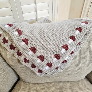 May include: A light gray crocheted blanket with a repeating pattern of maroon and white mushroom designs along the border. The blanket is folded over a light gray cushion, showcasing the textured stitch work.