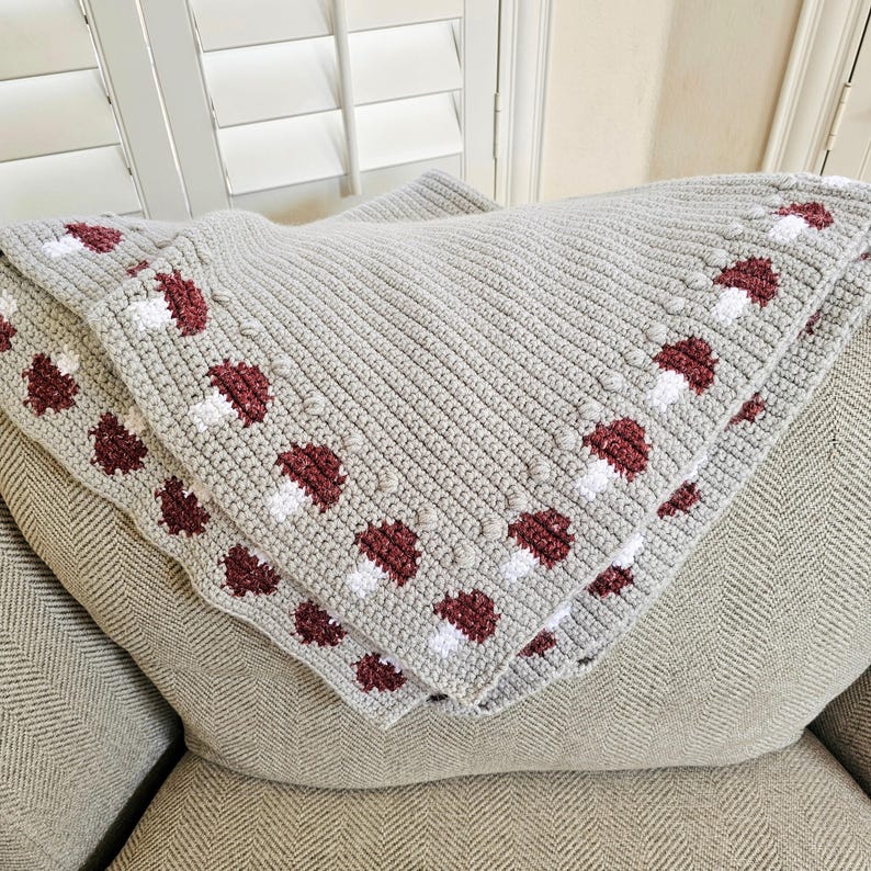 May include: A light gray crocheted blanket with a repeating pattern of maroon and white mushroom designs along the border. The blanket is folded over a light gray cushion, showcasing the textured stitch work.