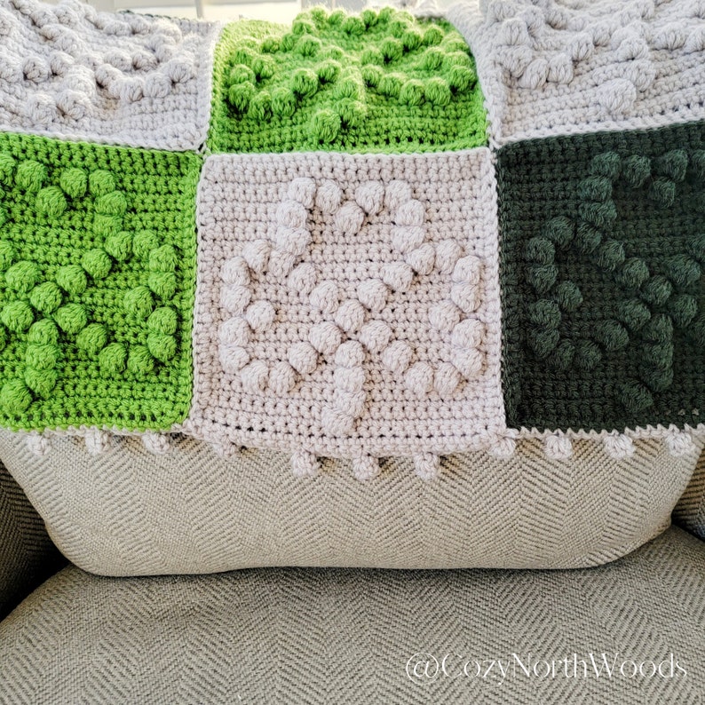 May include: A crocheted blanket with squares in shades of green and beige. The squares have a textured, puffy design.