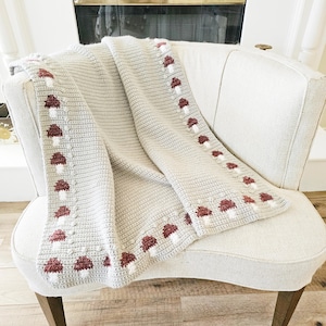 May include: A light gray crochet blanket with a repeating pattern of red and white mushrooms along the edges. The blanket is draped over a cream-colored armchair, showcasing its texture and design. The blanket is made of soft yarn.