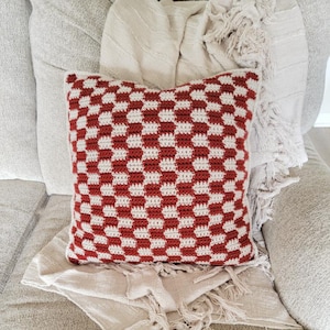 Puede incluir: Una almohada de crochet a cuadros rojos y blancos con ribete de flecos. La almohada est&aacute; sentada en un sof&aacute; marr&oacute;n claro con una manta blanca.