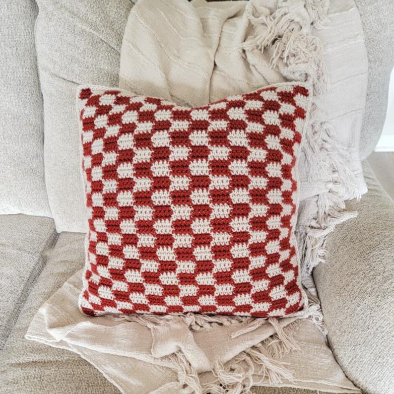 Puede incluir: Una almohada de crochet a cuadros rojos y blancos con ribete de flecos. La almohada est&aacute; sentada en un sof&aacute; marr&oacute;n claro con una manta blanca.