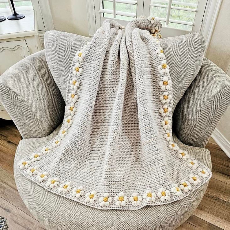 May include: A gray crocheted throw blanket with a white and yellow daisy border. The blanket is draped over a gray armchair.