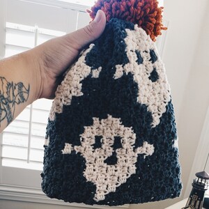 May include: A crocheted hat with a dark blue and white pattern and a large orange pom-pom on top.