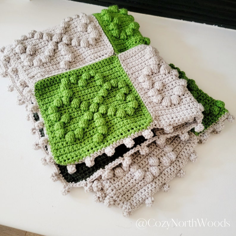 May include: A crocheted baby blanket with a patchwork design featuring squares of green and beige yarn. The blanket has a textured, puffy surface and is folded in thirds.