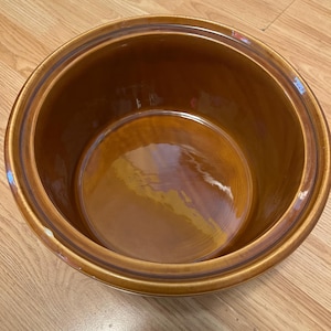 May include: A round, deep ceramic bowl with a glossy, smooth finish in a warm amber-brown color. The bowl features a wide, flat rim and a consistent, reflective glaze throughout the interior and exterior, showcasing a simple, functional design.