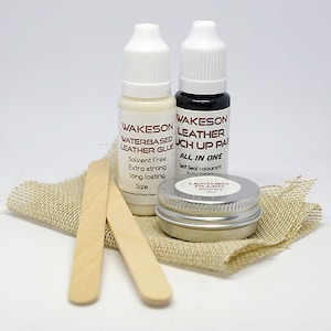 Op de afbeelding: Een lederreparatieset met twee flessen Wakeson Leather-producten, een kleine metalen doos en twee houten applicators. De flessen zijn gelabeld met "Waterbased Leather Glue" en "Touch Up Paint". De set ligt op een stuk jute.