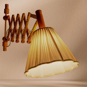 Peut inclure: Une applique murale dotée d'un bras articulé en accordéon en bois clair et d'un abat-jour en tissu plissé de couleur claire avec un bord festonné. La lampe est fabriquée en bois brun clair et comprend un cordon d'alimentation blanc.