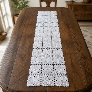 Könnte beinhalten: Ein weißer, rechteckiger Tischläufer mit einem sich wiederholenden Häkelmuster aus miteinander verbundenen Blumenmotiven und gewellten Rändern. Der Läufer besteht aus mehreren quadratischen Abschnitten, die zu einem langen Dekostück zusammengefügt sind.