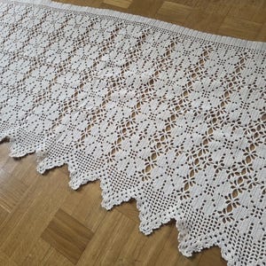 May include: Cream-coloured, open-weave crochet lace runner with a scalloped edge. The intricate floral pattern covers the entire length of the runner. The lace is displayed on a wooden surface.