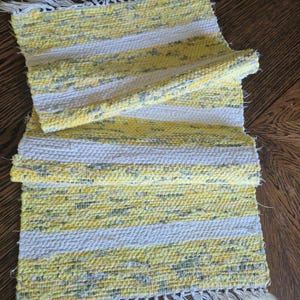 May include: Handwoven rectangular rug with yellow, white, and gray stripes. The rug has a fringed edge and is partially rolled up, showcasing its texture and design. A decorative textile for home decor.