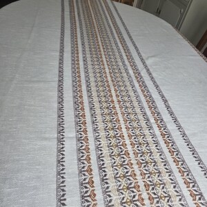 May include: A light-coloured oval tablecloth with a central patterned stripe. The stripe features repeating geometric designs in shades of brown, beige, and dark grey. The tablecloth is made of a woven fabric.
