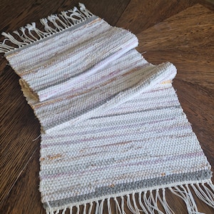 Puede incluir: Un camino de mesa rectangular tejido a mano con un borde con flecos. El camino de mesa presenta un patrón de rayas en tonos de blanco, gris y rosa y naranja apagados. El textil está hecho de tela tejida.