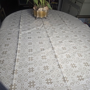May include: A large, oval, off-white tablecloth with a repeating pattern of light brown hash marks and geometric shapes. The tablecloth is draped over a table, with a decorative floral arrangement in a gold-colored container in the center.