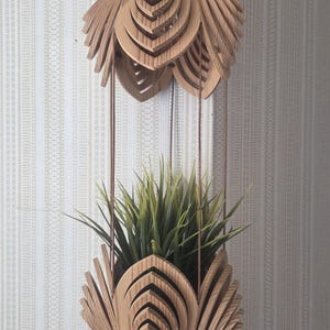 May include: A hanging wooden planter with a layered, floral-inspired design. The planter is suspended by thin brown cords and holds green grass-like plants. The wood has a natural, light brown color. The background is a patterned, off-white wall.