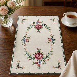 May include: Cream-colored rectangular table runner with a floral cross-stitch design. The embroidery features a central rose motif surrounded by a garland of green leaves and smaller floral accents in pink and blue.