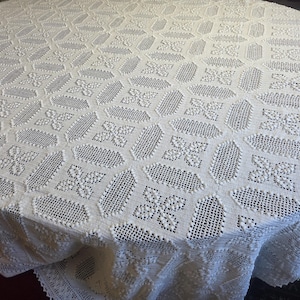 May include: A white, round tablecloth with a detailed, openwork design. The pattern features repeating floral and geometric shapes, creating a textured surface. The tablecloth is draped over a round table, showcasing its intricate craftsmanship.