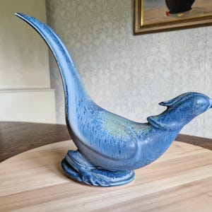 Może przedstawiać: Niebieska ceramiczna figurka ptaka z długą, zakrzywioną szyją i szczegółową głową. Ptak siedzi na podstawie, a szkliwo ma nakrapiany wygląd z odcieniami niebieskiego i zielonego. Figurka jest prezentowana na drewnianej powierzchni.