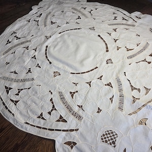 May include: A white, intricately embroidered tablecloth with a floral design. The tablecloth features cut-out patterns and scalloped edges, creating a delicate and elegant appearance. The design includes a central oval shape and detailed floral motifs.