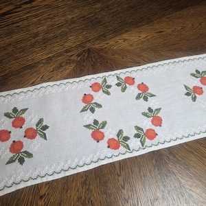 May include: White embroidered table runner with a repeating pattern of orange fruit and green leaves. The runner has a scalloped edge with green stitching. The fruit is arranged in a circular pattern in the center and in clusters on each end.