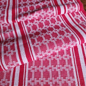 Op de afbeelding: Close-up van een rood en wit geweven textiel. De stof heeft een herhalend patroon van vierkanten en rechthoeken, met verticale strepen. Het kleurenschema is een levendig rood en wit, wat een visueel aantrekkelijk contrast creëert. Het textiel lijkt gevouwen.