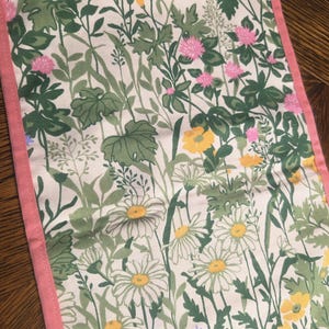 May include: A rectangular table runner with a floral pattern. The design features white daisies, yellow flowers, pink clover, and green foliage on a cream background. The edges are trimmed with a pink border.