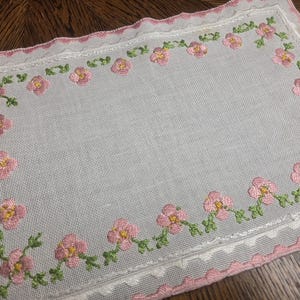 以下が含まれることがあります： ピンクの花柄の縁取りが施された、白い長方形の刺繍入りドイリー。 ドイリーには、黄色の中心と緑の葉を持つピンクの花が描かれています。 エッジはピンクのスカラップトリムで仕上げられています。