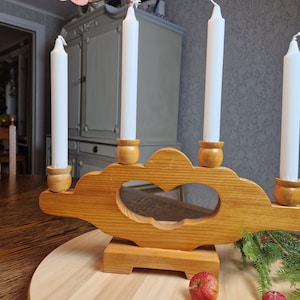 May include: A wooden candle holder with four white candles. The holder is light brown with a heart-shaped cutout in the center. The candles are tall and slender. The holder sits on a wooden base.