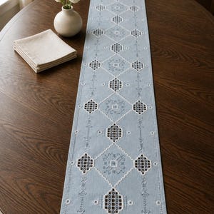 May include: A light blue embroidered table runner with a geometric pattern, displayed on a dark wood table. The runner features white and dark blue diamond-shaped designs. A small vase with white flowers and a stack of beige napkins are also on the table.