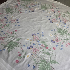 Könnte beinhalten: Ein hellgraues Tischtuch mit floralem Muster. Das Design zeigt Sträuße aus weißen Gänseblümchen, rosa und roten Blumen, blauen Glockenblumen und grünen Farnen. Das Tischtuch ist aus einem weichen Stoff, möglicherweise Leinen.