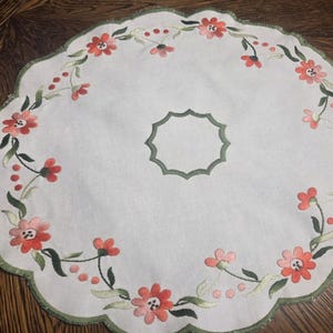 May include: White, scalloped-edge placemat with embroidered floral design. The embroidery features red and pink flowers, green leaves, and red berries. A green border and a central green geometric shape complete the design.