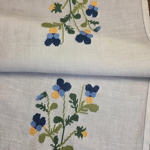 Può includere: Due tovaglioli di lino azzurro con ricami floreali. Ogni tovagliolo presenta un motivo a punto croce di fiori e foglie blu, gialli e verdi. I bordi sono rifiniti con un bordo cucito blu.