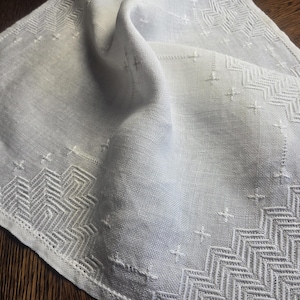 Peut inclure: Une serviette de table en lin blanc avec des motifs géométriques brodés complexes sur les bords. La broderie présente des motifs en zigzag et en point de croix. La serviette est pliée, révélant la texture du tissu.