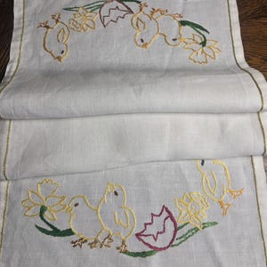 May include: White linen table runner with embroidered Easter-themed design. Features yellow chicks, daffodils, and eggshell details. The edges are trimmed with a gold border. A decorative piece for the dining table.