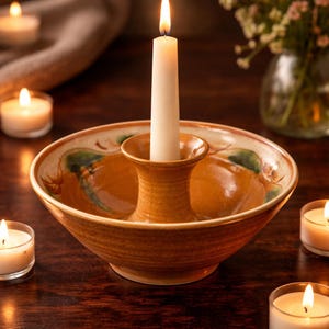 Peut inclure: Un bougeoir en céramique avec un pilier central pour une longue bougie blanche. Le bougeoir a une forme de bol avec un glaçage marron et orange et des motifs de feuilles vertes. Plusieurs bougies chauffe-plat allumées sont éparpillées.