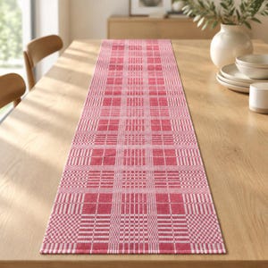Puede incluir: Un camino de mesa rectangular con un patrón de cuadros tejido en tonos rojos y blancos. El tejido presenta un diseño de cuadrícula repetitivo con bloques rojos sólidos que se alternan con secciones de rayas blancas.