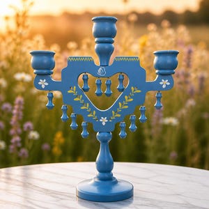 Peut inclure: Un candélabre en bois bleu avec une découpe centrale en forme de cœur, trois bougeoirs et des pendentifs décoratifs suspendus. L'objet est orné de motifs peints à la main de feuilles jaunes et de fleurs blanches, avec un petit logo cygne blanc.