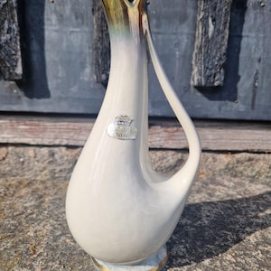 May include: A white ceramic pitcher with a gold-colored rim and handle. The pitcher has a unique, curved design and a small emblem on the front. The base has a gold trim. The pitcher is approximately 10 inches tall.