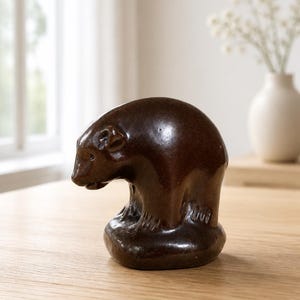Pode incluir: Uma estatueta de urso de cerâmica marrom. O urso é retratado em posição agachada, com a cabeça baixa. A estatueta tem um acabamento liso e brilhante e está sobre uma mesa de madeira. Um vaso com flores brancas está ao fundo.